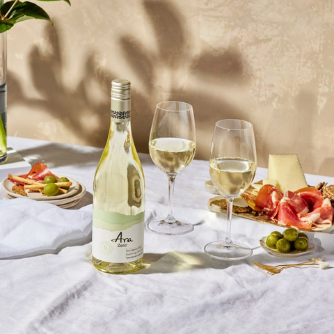 Butelka wina bezalkoholowego Guiltfree Ara Zero Sauvignon Blanc z kieliszkami i przekąskami na białym obrusie.