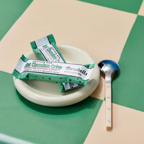 Baton proteinowy Guiltfree Barebells Mint Chocolate Crisp na małym talerzyku z łyżeczką na zielono-beżowej szachownicy