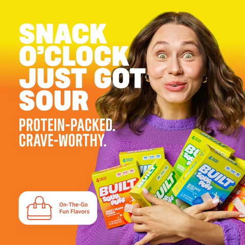 Kobieta w fioletowym swetrze trzyma opakowania batonów proteinowych Guiltfree Built Sour Puff o smaku brzoskwiniowym