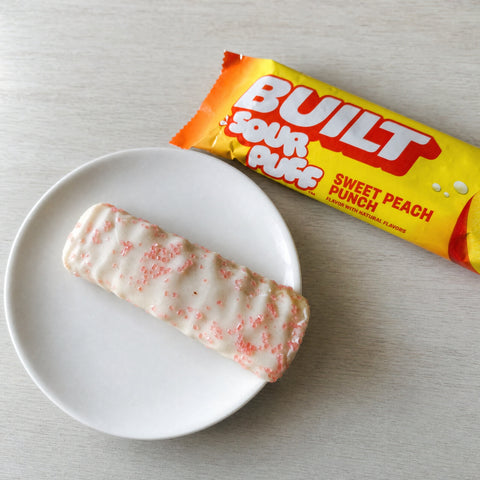 Guiltfree baton proteinowy Built Sour Puff Sweet Peach Punch z jasną polewą i różowymi kryształkami na talerzyku