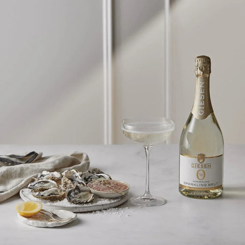 Butelka bezalkoholowego wina musującego Guiltfree Giesen Sparkling Brut obok kieliszka i talerza świeżych ostryg.