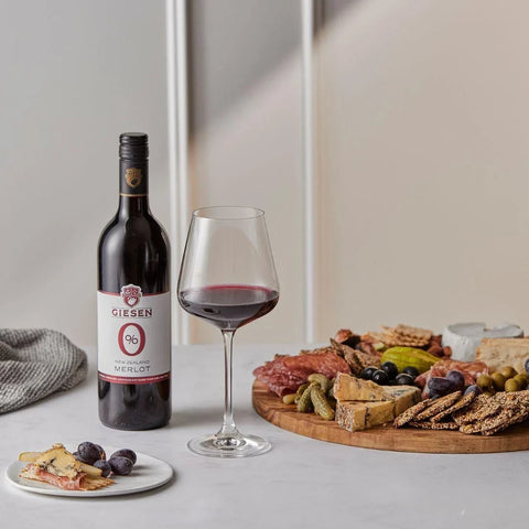 Butelka wina bezalkoholowego Guiltfree Giesen 0% Merlot z kieliszkiem i deską serów oraz przekąsek na stole