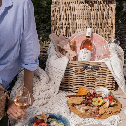 Butelka wina Guiltfree Giesen 0% Rosé w wiklinowym koszu piknikowym z deską serów i owoców na zewnątrz.