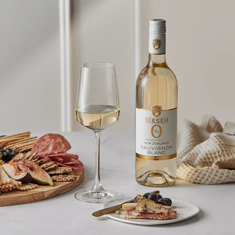 Butelka Guiltfree Giesen 0% Sauvignon Blanc obok kieliszka wina i deski z przekąskami, serami oraz owocami.