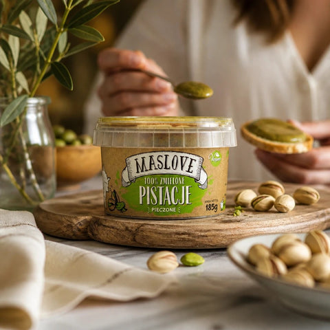 Słoik masła z pieczonych pistacji Maslove na drewnianej desce w ofercie Guiltfree, obok rozsypane orzechy pistacjowe.