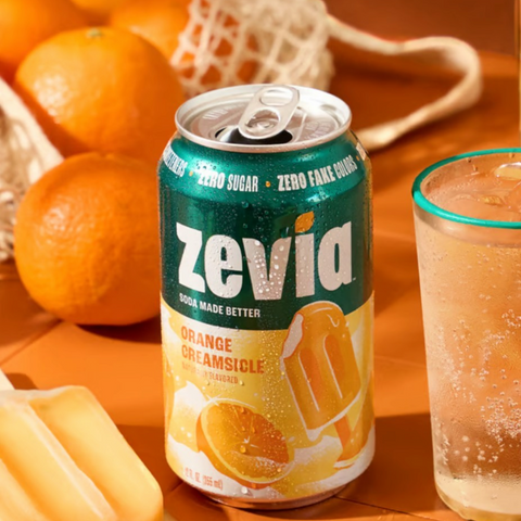 Puszka napoju gazowanego Zevia Orange Creamsicle bez cukru, obok świeżych pomarańczy i lodów na patyku.
